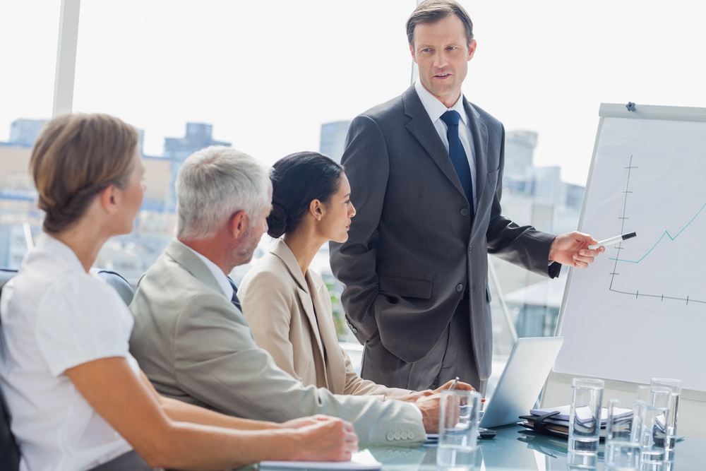 Businessman pointing at whiteboard during a meeting in front of attentive colleagues Businessman pointing at whiteboard during a meeting in front of attentive colleagues